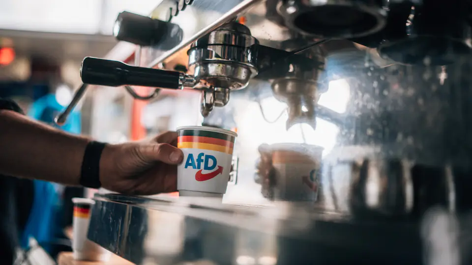 Eine Hand stellt einen Pappbecher mit AfD-Logo unter den Auslauf einer Siebträger-Kaffeemaschine.