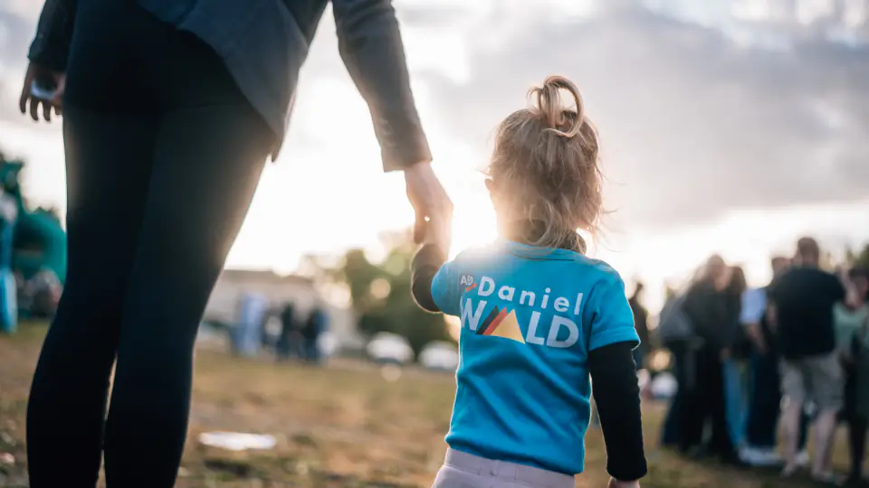 Ein Kind in einem blauen T-Shirt mit dem Aufdruck „Daniel Wald" geht im Gegenlicht an der Hand eines Erwachsenen über die Festwiese.