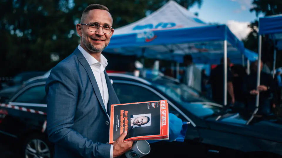 Daniel Wald steht mit einer Zeitschrift in der Hand vor den blauen Pavillons des Infostands und lächelt in die Kamera.