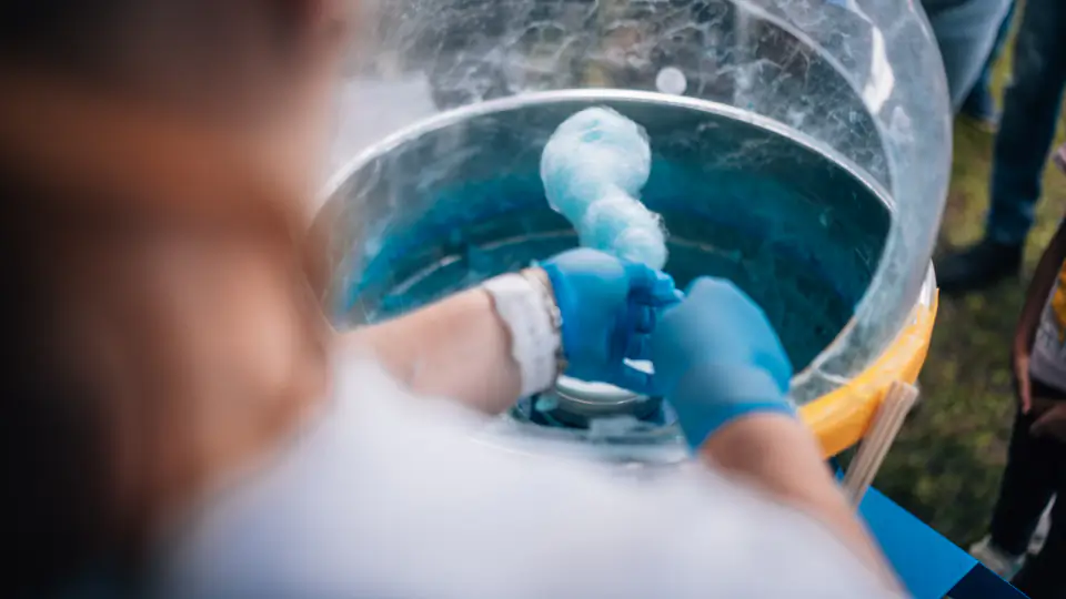 Hände in blauen Handschuhen drehen an einer Zuckerwattemaschine hellblaue Zuckerwatte auf einen Stab.